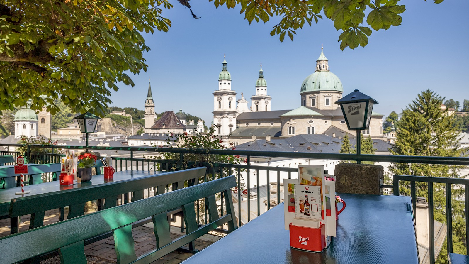 This atmospheric image captures a romantic beer garden terrace at sunset. Long wooden benches sit beneath a leafy canopy adorned with decorative lanterns, creating warm light and a cozy ambiance. In the foreground, two people toast with beer glasses, while other guests relax and chat in the background. The setting sun casts long shadows on the ground and bathes the baroque cityscape—with its domes and church towers—in golden light. The scene conveys a sense of tranquility, togetherness, and the charm of a summer evening in the historic old town of Salzburg.