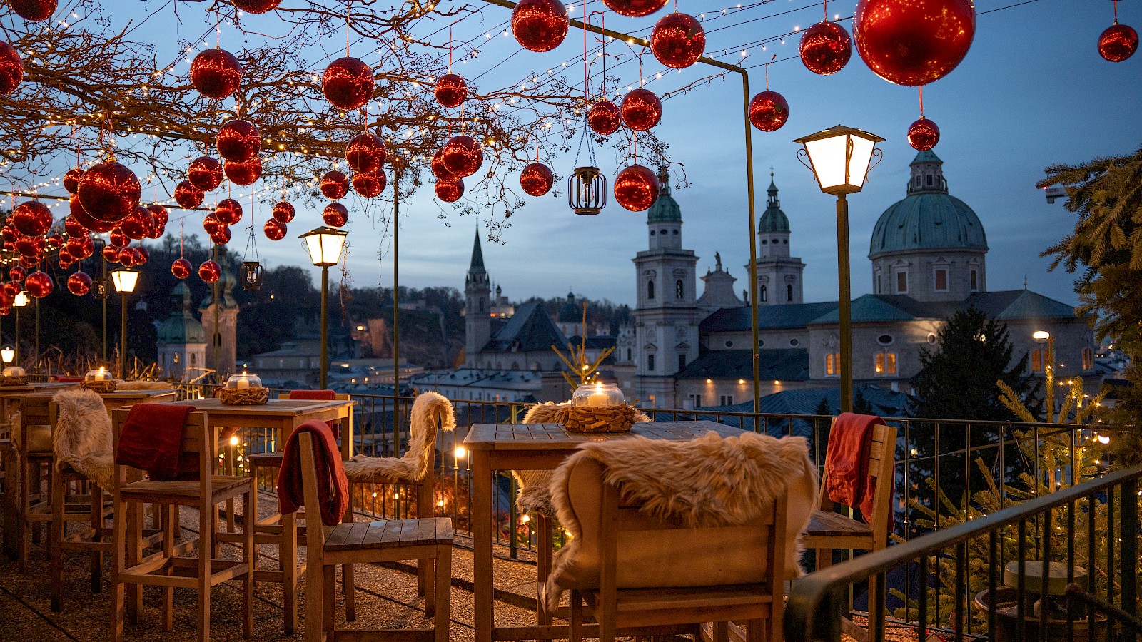 Diese stimmungsvolle Aufnahme zeigt eine romantische Biergarten-Terrasse bei Sonnenuntergang. Die langen Holzbänke stehen unter einem Blätterdach mit dekorativen Lampions, das warmes Licht und eine gemütliche Atmosphäre schafft. Im Vordergrund stoßen zwei Personen mit Biergläsern an, während im Hintergrund weitere Gäste entspannt plaudern. Die untergehende Sonne wirft lange Schatten auf den Boden und taucht die barocke Stadtkulisse mit Kuppeln und Kirchtürmen in goldenes Licht. Die Szene vermittelt Ruhe, Geselligkeit und den Zauber eines Sommerabends in der historischen Altstadt von Salzburg.
